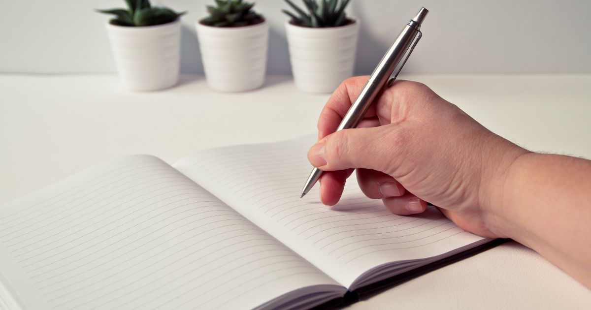 A person writing in a notebook with pens.